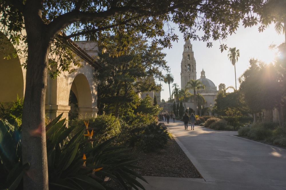 A garden path lined with trees and Spanish colonial style buildings in a large park